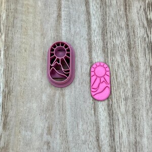 May include: A pink, oval-shaped cookie cutter and a pink cookie. The cutter features a sun and mountain design. The cookie has the same design. The cutter is approximately 7.6 cm long.