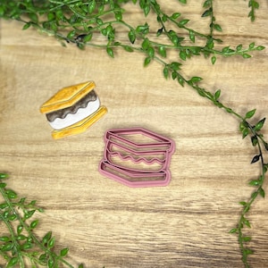 May include: Two cookie cutters in the shape of a s'more. The top cutter is yellow and brown, and the bottom cutter is pink.