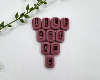 Puede incluir: Once cortadores de galletas rectangulares de color malva con varios diseños interiores están dispuestos sobre una superficie blanca. Una ramita de follaje verde se encuentra en la esquina superior izquierda. Los diseños incluyen rectángulos, óvalos y líneas.