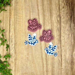 May include: Three cookie cutters in the shape of a bird with floral details. Two are pink and one is white with blue floral details.