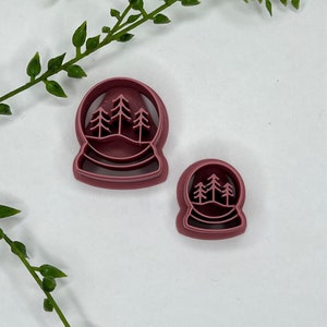 May include: Two red plastic cookie cutters shaped like snow globes with three trees inside. The larger cutter is about 2 inches in diameter and the smaller cutter is about 1 inch in diameter.