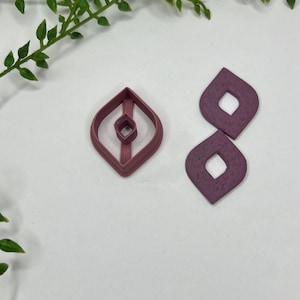 May include: Two pink polymer clay earrings in the shape of a diamond with a smaller diamond cut out in the center. The earrings are next to a cookie cutter in the shape of a diamond with a smaller diamond cut out in the center.