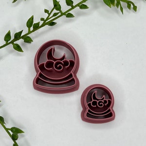May include: Two pink plastic cookie cutters shaped like snow globes with a crescent moon and clouds inside. The larger cutter is in the foreground and the smaller cutter is in the background.