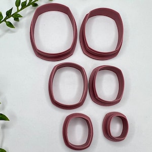 May include: Set of six plastic cookie cutters in various sizes. The cutters are shaped like rounded rectangles and are a dusty rose color.