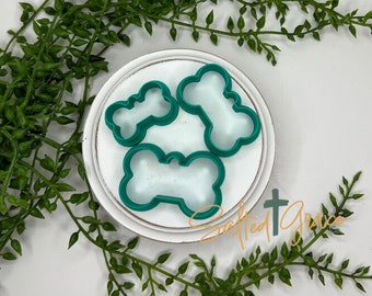 Puede incluir: Tres cortadores de galletas de plástico verde en forma de hueso en un plato blanco. El plato está sobre una superficie blanca.