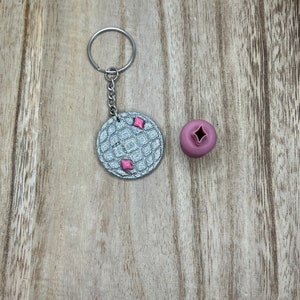 May include: A silver keyring with a waffle pattern and pink accents. The keyring is attached to a silver key ring. A pink waffle-shaped charm is also included.