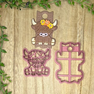 May include: Three pink plastic cookie cutters shaped like a Highland cow. The cow is wearing a flower crown.