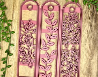 May include: Three pink plastic cookie cutters shaped like rectangular bookmarks. The leftmost cutter features a floral design, the middle cutter features a leafy design, and the rightmost cutter features a floral design.