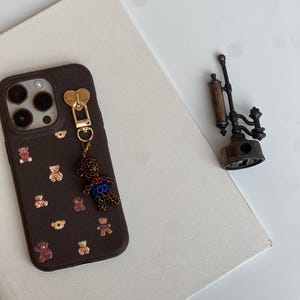 May include: A brown phone case decorated with small teddy bear motifs. A gold heart-shaped charm and a teddy bear charm hang from the case. A vintage-style metal object is placed nearby on a white surface.