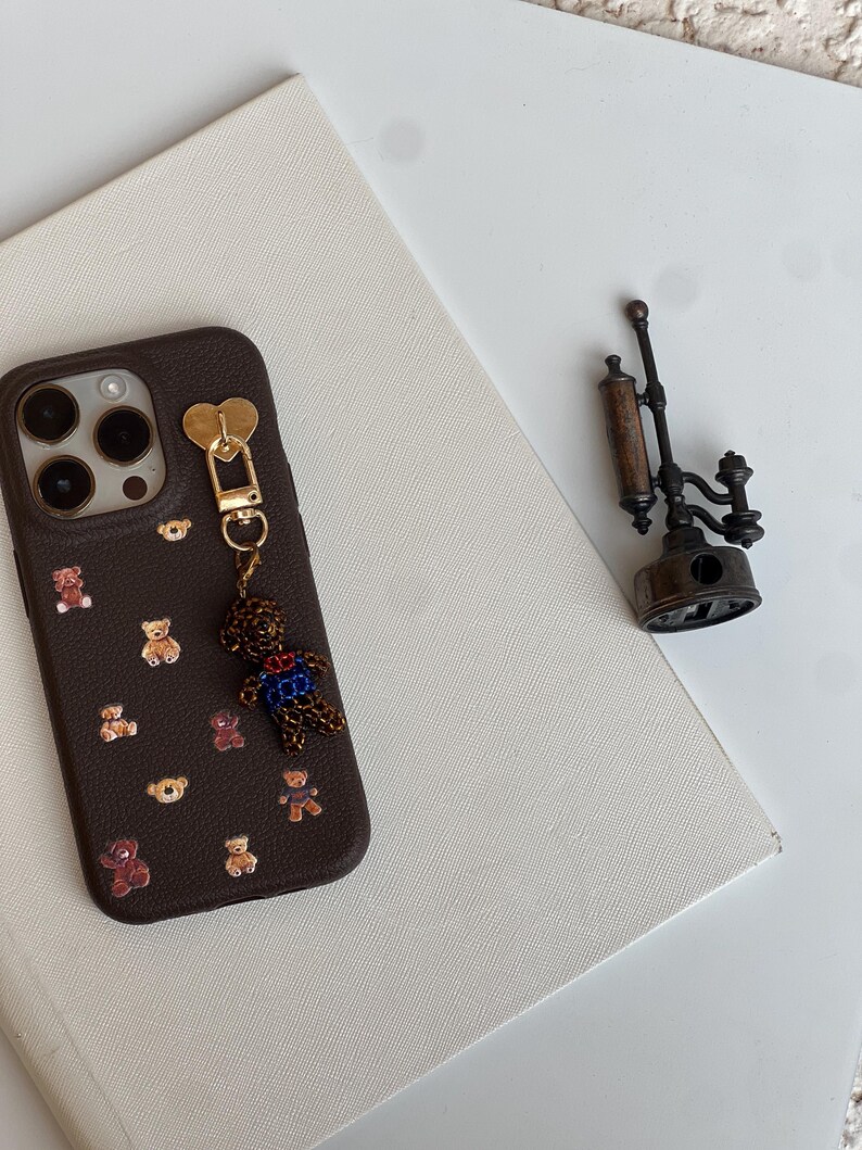 May include: A brown phone case decorated with small teddy bear motifs. A gold heart-shaped charm and a teddy bear charm hang from the case. A vintage-style metal object is placed nearby on a white surface.