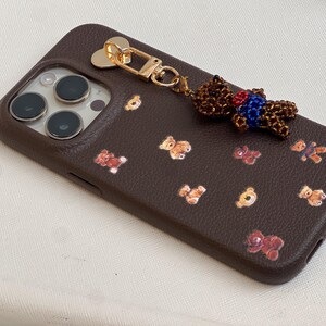 May include: A brown phone case featuring a repeating pattern of teddy bear illustrations. A beaded teddy bear charm with a gold-coloured clasp is attached. The case has a textured finish.