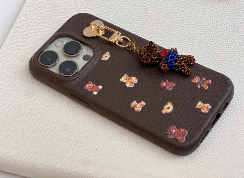 May include: A brown phone case featuring a repeating pattern of teddy bear illustrations. A beaded teddy bear charm with a gold-coloured clasp is attached. The case has a textured finish.