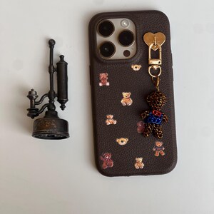 May include: A brown phone case adorned with teddy bear motifs and a dangling teddy bear charm. The case features a gold heart-shaped clasp and a dark brown leather texture. A vintage metal lamp is also visible.