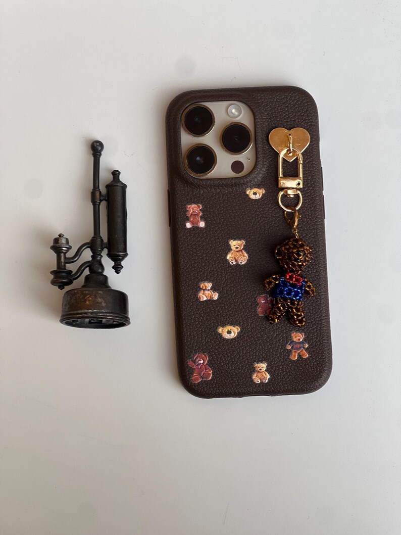May include: A brown phone case adorned with teddy bear motifs and a dangling teddy bear charm. The case features a gold heart-shaped clasp and a dark brown leather texture. A vintage metal lamp is also visible.