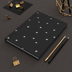 May include: A black notebook with a white heart pattern on the cover. The notebook is lying on a dark gray surface with two pencils and a gold metal decorative stand.