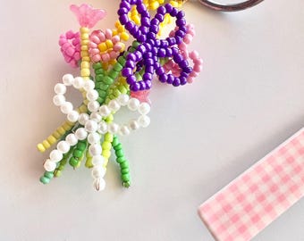 May include: A beaded flower bouquet keychain with pink, yellow, green, white, and purple beads. The keychain is attached to a set of car keys. A pink and white checkered object is in the background.