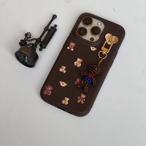 May include: A brown phone case adorned with small teddy bear decorations. A beaded teddy bear charm is attached to the case. A small, antique-style metal object is to the left of the phone case.