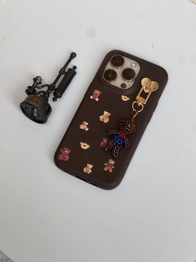May include: A brown phone case adorned with small teddy bear decorations. A beaded teddy bear charm is attached to the case. A small, antique-style metal object is to the left of the phone case.