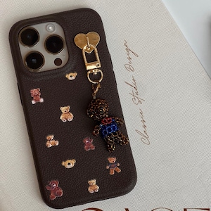 May include: A dark brown phone case decorated with miniature teddy bear designs. A beaded teddy bear charm is attached. The words "PHONE" and "CASE" are at the top and bottom, and "Classic Studio Design" is written on the side.
