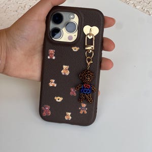 May include: A brown phone case decorated with small teddy bear decals. A charm of a teddy bear with a blue and red outfit hangs from the case. The case has a gold heart-shaped attachment.