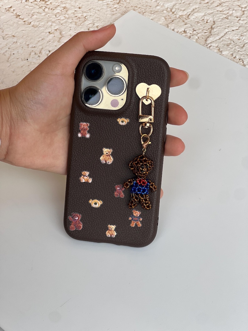 May include: A brown phone case decorated with small teddy bear decals. A charm of a teddy bear with a blue and red outfit hangs from the case. The case has a gold heart-shaped attachment.