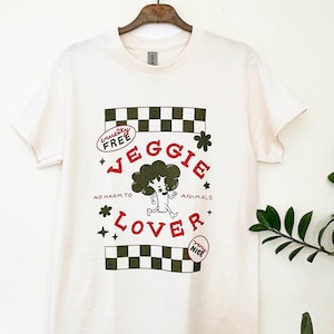 Op de afbeelding: Crèmekleurig T-shirt met een grafisch ontwerp met een cartoon broccoli karakter en de woorden "Veggie Lover". Het ontwerp bevat de tekst "Cruelty Free" en "No Harm to Animals" in rood en groen.