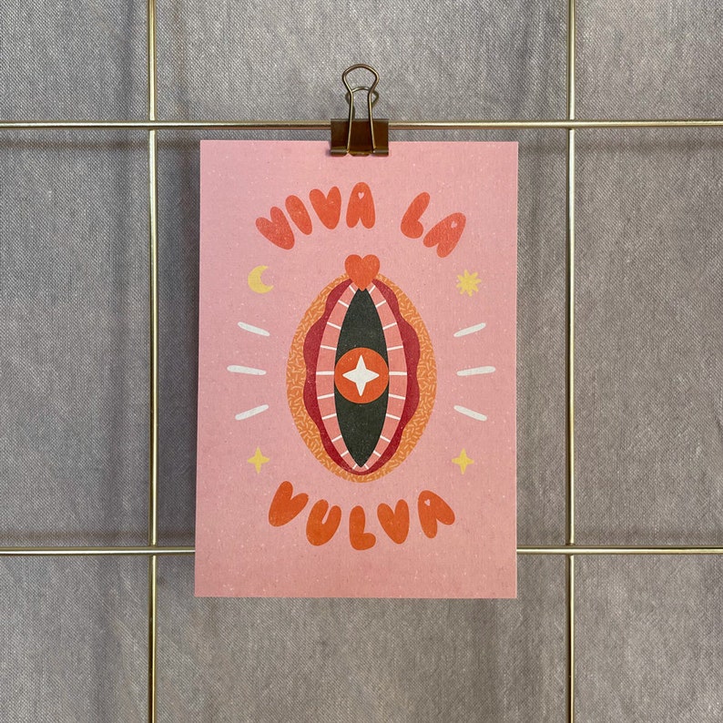 以下が含まれることがあります： オレンジ色の文字で「VIVA LA VULVA」と書かれたピンク色のプリント。イラストは、ハート、星、その他の装飾要素を備えた、様式化された外陰部のデザインを特徴としています。プリントは、金色のワイヤーグリッド上のクリップで保持されています。