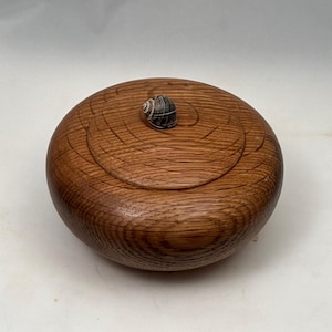 May include: A round, wooden box with a lid, featuring a natural wood grain finish. A small, dark-colored seashell is affixed to the top of the lid. The box is designed with a circular pattern on the lid.