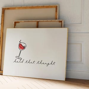 Könnte beinhalten: Ein weißer Leinwanddruck mit einer Weinglas-Illustration und dem Text "hold that thought". Das Kunstwerk lehnt an einer Wand mit zwei weiteren gerahmten Leinwänden. Der Raum hat einen Holzboden und weiße Wände.