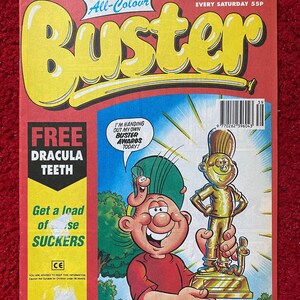 Op de afbeelding: Een vintage comicboekomslag met een cartoonfiguur dat een gouden trofee vasthoudt. De omslag is rood en geel met de titel "Buster" in grote, vetgedrukte letters. De tekst "Free Dracula Teeth" en "Get a load of these suckers" is ook zichtbaar.
