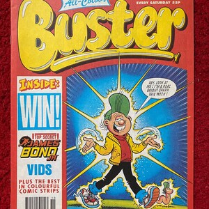 Pode incluir: Uma capa de banda desenhada para a revista Buster, com uma personagem de banda desenhada com um chapéu verde e uma jaqueta amarela. O texto da capa diz "All-Colour Buster" e "Inside: Win! Top Secret James Bond Jr. Vids Plus the Best in Colourful Comic Strips".