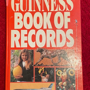 Op de afbeelding: Een rood hardback boek met de titel "Guinness Book of Records" in witte tekst. De omslag toont verschillende afbeeldingen, waaronder een jong meisje dat een grote citroen vasthoudt, een cheeta-beeld, een persoon die vuur spuit en een sterke man die gewichten tilt. Het boek is uit de editie van 1980.