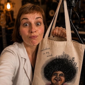 Puede incluir: Una bolsa de tela beige con una ilustración impresa de una persona con un peinado afro grande y oscuro, vistiendo una camisa de lunares rojos. El texto "melodies in my mind" está impreso sobre la ilustración, junto a notas musicales.