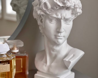 May include: White plaster bust of a man with short curly hair. The bust is sitting on a gray surface next to a bottle of perfume and a gold chain necklace with white pearls.