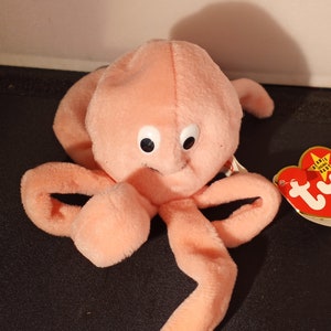 May include: A plush, peach-colored toy octopus with large, round, black-pupiled eyes. The octopus has eight long, curved tentacles. A red and yellow tag is attached to the toy.