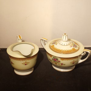 Può includere: Servizio di lattiera e zuccheriera coperte in porcellana color crema. Il set presenta finiture dorate e decorazioni floreali. Lo zuccheriera ha un coperchio con un pomello decorativo. La lattiera ha un manico e un beccuccio.