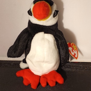 May include: A plush puffin toy with a black body, white belly, and red feet. The puffin has a large orange beak, black eyes, and a TY tag attached. The toy is approximately 6 inches tall.
