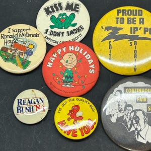 Op de afbeelding: Een verzameling vintage knopen met verschillende ontwerpen, waaronder een kikker, een cartoonfiguur, een politieke campagne en een insect. De knopen zijn kleurrijk en hebben verschillende slogans en afbeeldingen.