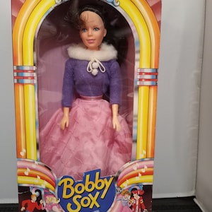 May include: A vintage Bobby Sox fashion doll, Norma-Jean, in its original packaging. The doll is dressed in a purple sweater, pink skirt, and a white collar. The box has 1950s-themed graphics and text that reads "The Fun & Fabulous Fifties are Back!"
