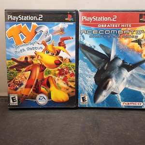 May include: Two PlayStation 2 video game cases. The first features a cartoon kangaroo with the title "TY the Tasmanian Tiger Bush Rescue." The second case is for "Ace Combat 04: Shattered Skies" with a jet fighter image.