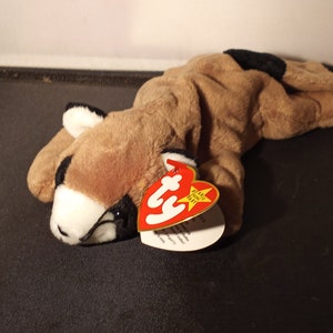 May include: A plush toy cougar in a lying position, featuring a tan body with black accents on the back and ears. The cougar has a white muzzle and black eyes. A red and white tag is attached to the toy.