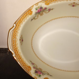 May include: Oval serving dish with a cream-colored interior and a decorative gold border. The border features floral and scroll designs with pink and purple flowers. The dish has gold handles and a delicate, patterned edge.
