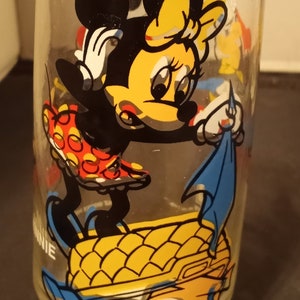 May include: Clear glass with a cartoon illustration of Minnie Mouse. Minnie is wearing a red and yellow polka-dot dress, black shoes, and a yellow bow. The glass has a yellow and blue design at the base.
