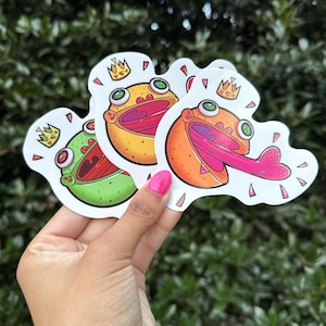 May include: Three frog stickers with crowns and open mouths. The frogs are green, orange, and yellow. The stickers are on a white background.