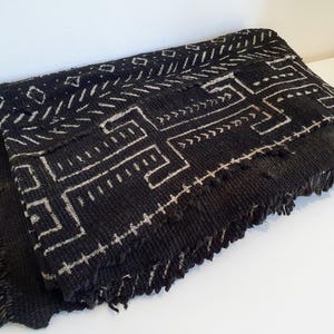 Könnte beinhalten: Gefaltetes schwarzes Textil mit weißem geometrischen Muster. Der Stoff hat eine gewebte Textur und einen Fransenrand. Das Design zeigt lineare und eckige Formen, die einen auffälligen Kontrast erzeugen.