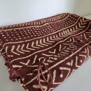 May include: A folded textile featuring a deep maroon background with a contrasting cream-coloured geometric pattern. The design includes repeating chevron stripes, rows of small dots, and diamond shapes, finished with frayed edges along the side.