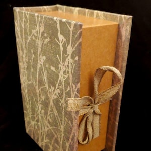 May include: A small, rectangular box with a brown paper exterior and a decorative, patterned cover. The cover features a light green background with a white floral design. A brown ribbon is tied around the box.