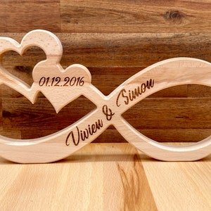 Könnte beinhalten: Ein Holz-Unendlichkeitszeichen mit zwei darin geschnitzten Herzen. Das Datum "01.12.2016" und die Namen "Vivien & Simon" sind in das Symbol graviert.