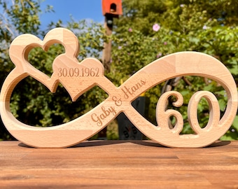 May include: A wooden infinity symbol with two hearts carved into the top. The symbol is engraved with the text "Gaby & Hans" and the date "30.09.1962". The number "60" is also engraved on the symbol.