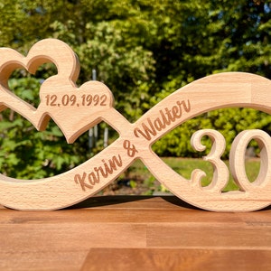 Könnte beinhalten: Ein Holz-Unendlichkeitszeichen mit zwei Herzen, die in die Oberseite geschnitzt sind. Das Symbol ist mit den Namen "Karin & Walter" und dem Datum "12.09.1992" graviert. Die Zahl "30" ist ebenfalls in das Symbol graviert.
